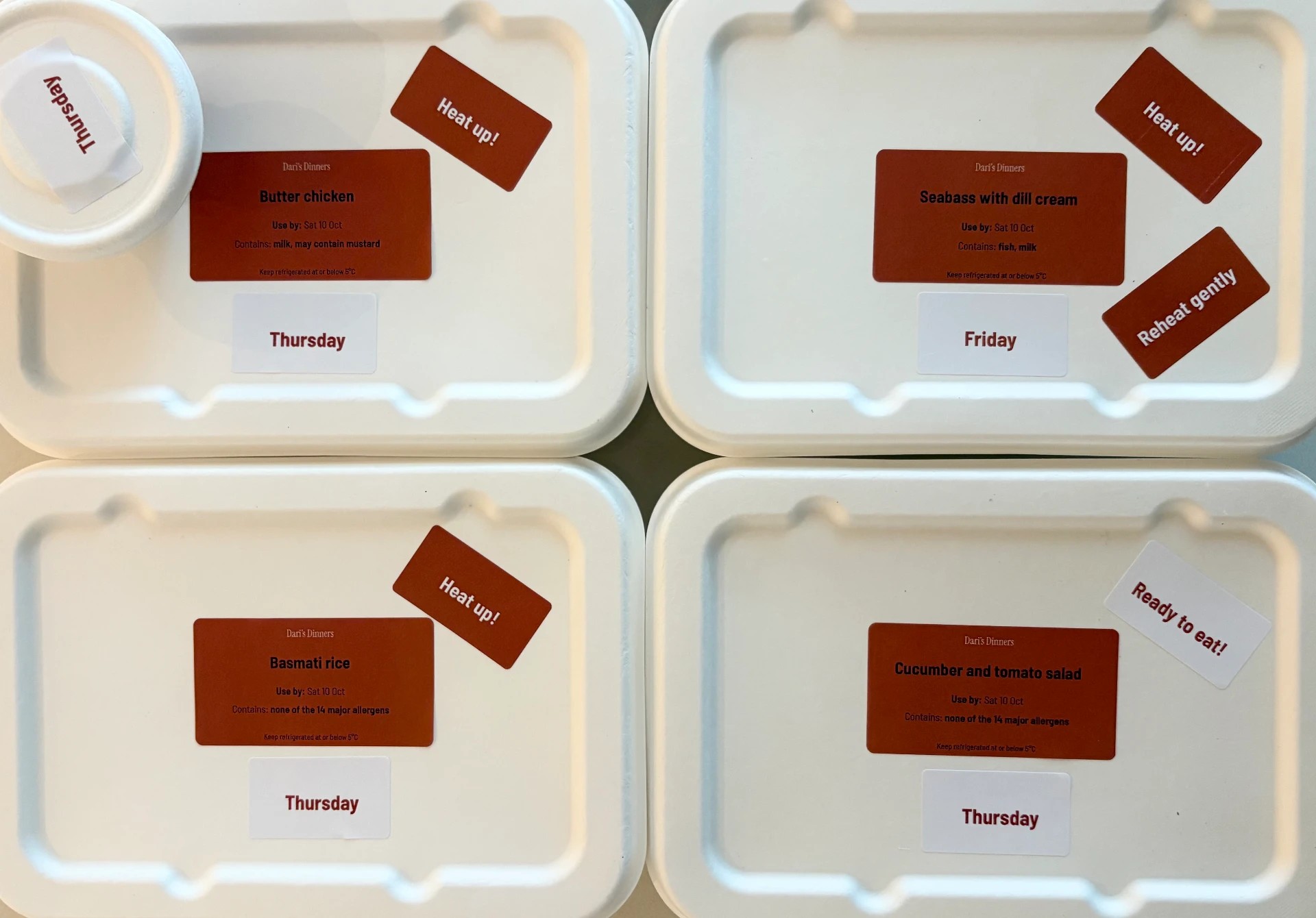 Four labelled containers seen from above: butter chicken, sea bass with dill cream, basmati rice and cucumber and tomato salad