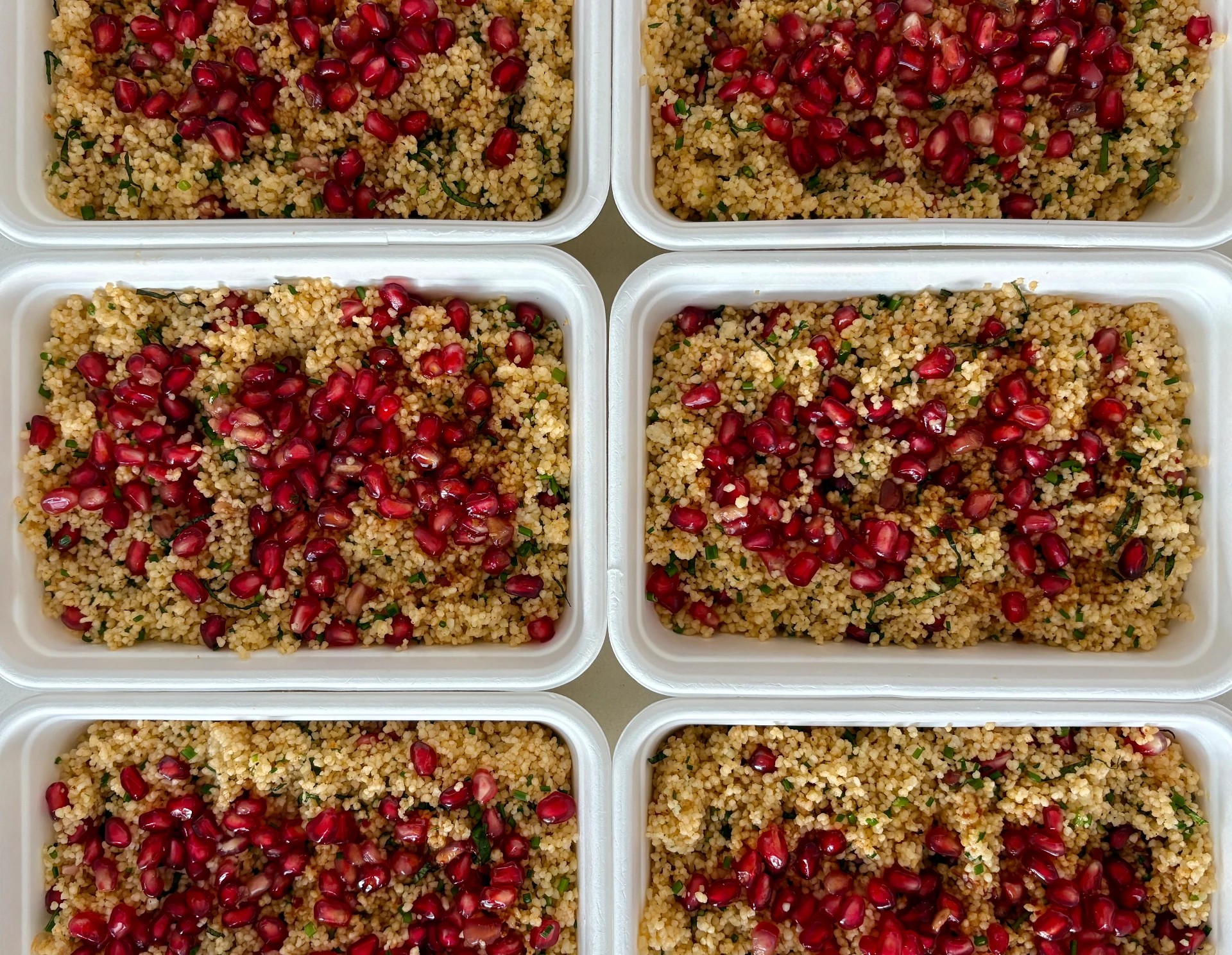 Six containers of herbed grain salad topped with pomegranate seeds, shot from above
